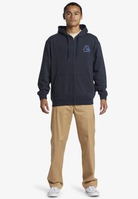 Mladý muž v námořníkovom zipsovom mikinovom svetri s logom vlny, béžových rovných nohaviciach a bielych teniskách, stojí pred bielym pozadím.