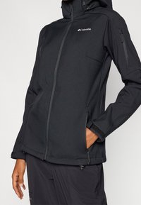 Czarna kurtka softshell z zamkiem błyskawicznym z przodu, regulowanym kapturem i bocznymi kieszeniami. Posiada subtelne logo oraz mankiety na rzepy zapewniające pewne dopasowanie.