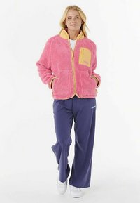 Giacca rosa pelosa con dettagli gialli e tasche, abbinata a pantaloni larghi blu navy. La giacca presenta un colletto alto e chiusura a zip.