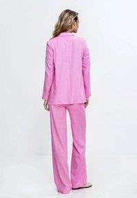 Dopasowana różowa marynarka i pasujące szerokie spodnie uszyte z lekkiego materiału. Marynarka ma wyłogowy kołnierz i gładką fakturę.