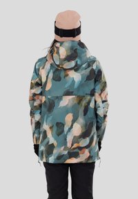 Osoba oblečená v kapucovém, barevném modro-zeleno-bežovém vzorovaném kabátu a růžové čepici s černým popruhem, stojící zády.