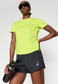 Neon keltainen urheilupaita, jossa on pieni harmaa Nike-logo; yhdistetty mustiin shortseihin, joissa on valkoinen Nike-swoosh; malli pitelee mustaa vesipulloa.