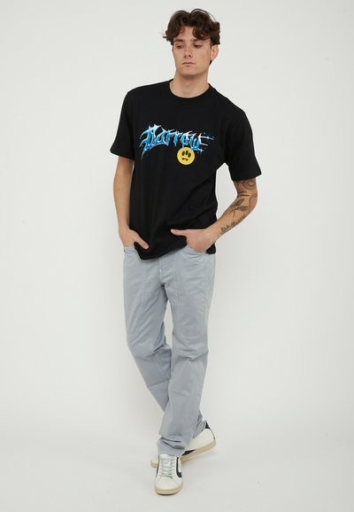 Young man with short curly hair wearing a black graphic T-shirt, light gray pants, white sneakers, and displaying a tattoo on his left forearm.