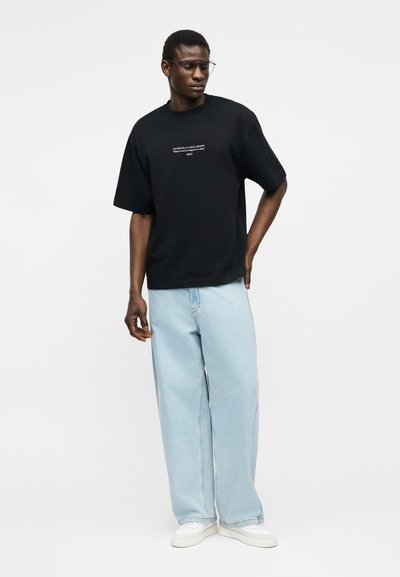 Άνδρας που φοράει μαύρο φαρδύ μπλουζάκι με λευκό κείμενο, ανοιχτό μπλε τζιν παντελόνι με φαρδιά μπατζάκια, λευκά αθλητικά παπούτσια και γυαλιά, στέκεται μπροστά σε απλό φωτεινό φόντο.