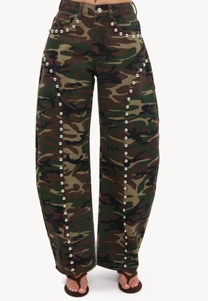 Pantalon camouflage à coupe ample avec des clous argentés soulignant les poches et les coutures latérales, porté avec des tongs marron.