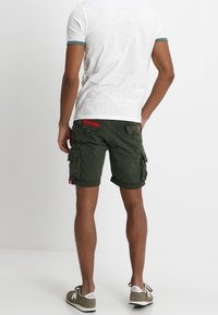 Vihreät cargo-shortsit, joissa on useita taskuja ja käännetyt lahkeet, yhdistettynä lyhythihaiseen valkoiseen paitaan, jossa on teal-värisiä yksityiskohtia, sekä rentoihin lenkkareihin.