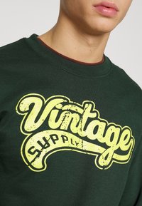 Tmavě zelená mikina s žlutým grafickým nápisem "Vintage Supply". Kulatý výstřih a měkká textura. Výrazné opotřebení na textovém designu.