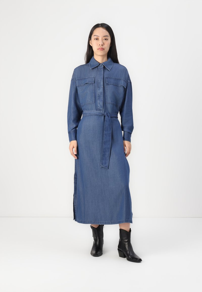 Robe chemise en denim avec manches longues, deux poches poitrine, une taille ceinturée et des fentes latérales. Couleur bleu uni avec une texture lisse.