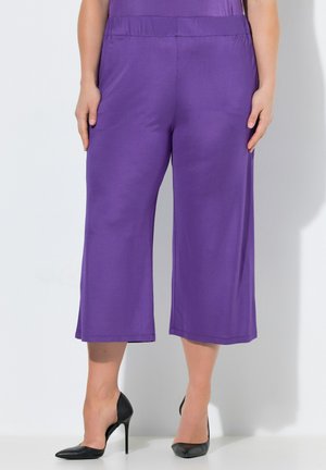 Kobieta w fioletowych szerokich spodniach do połowy łydki i czarnych, szpiczastych szpilkach stoi przy białej ścianie.