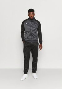 Trenirka v temno sivi barvi s črnimi poudarki, ki vključuje jakno z zadrgo in zožene hlače; narejena iz gladkega, lahkega materiala s športnim dizajnom.