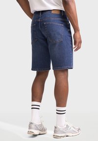 Bărbat purtând pantaloni scurți din denim albastru închis, șosete albe cu dungi negre și sneakerși albi New Balance, stând în fața unui fundal simplu.