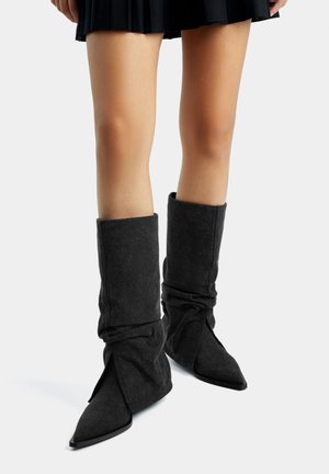 Gambe con indosso una gonna nera plissettata e stivali neri larghi a metà polpaccio con punte appuntite, su uno sfondo chiaro e uniforme.