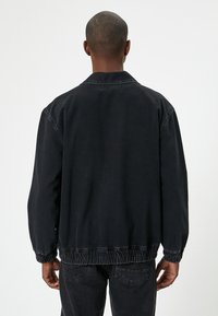 Hombre de espaldas que lleva una chaqueta negra suelta con puños y dobladillo elásticos, combinada con jeans de mezclilla negros, frente a un fondo blanco sencillo.