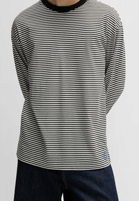 Gestreiftes Langarmhemd in Schwarz und Weiß mit einem gerippten schwarzen Kragen. Verfügt über ein blaues Logo auf der unteren linken Seite, kombiniert mit dunklen Jeans.