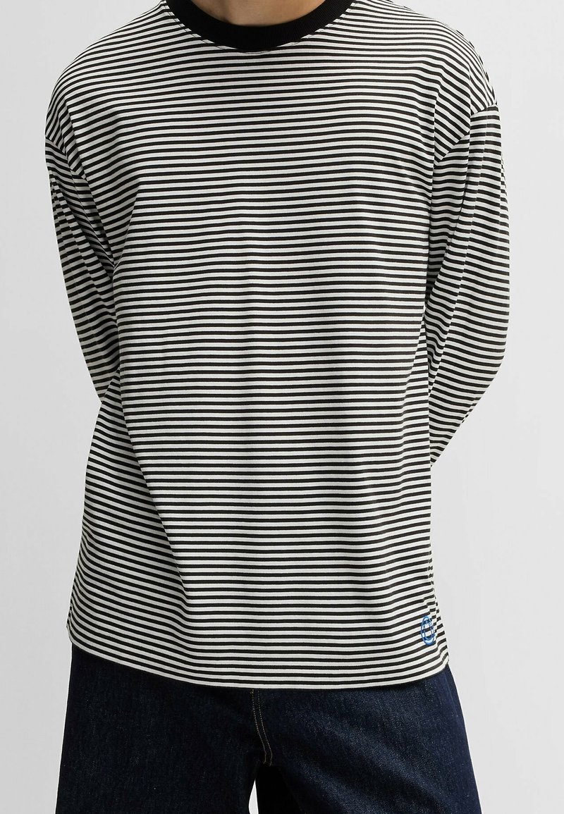 Gestreiftes Langarmhemd in Schwarz und Weiß mit einem gerippten schwarzen Kragen. Verfügt über ein blaues Logo auf der unteren linken Seite, kombiniert mit dunklen Jeans.