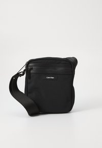 Borsa a tracolla nera realizzata in tessuto voluminoso con tracolla regolabile, dotata di chiusura superiore a zip e tasca con zip frontale. Logo Calvin Klein in evidenza.