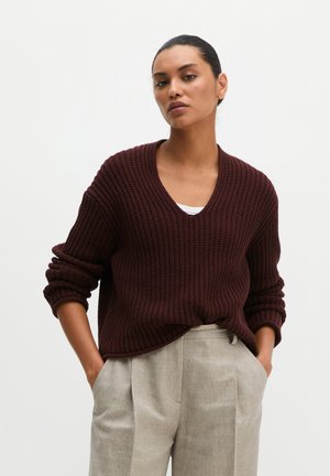 Femme aux cheveux foncés portant un pull en maille bordeaux et un pantalon plissé gris clair, debout, les mains dans les poches, devant un fond uni.