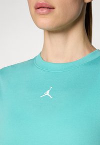 Camiseta turquesa con cuello redondo y un logo de Jordan blanco en el pecho. La tela parece suave y tiene una textura ligera.