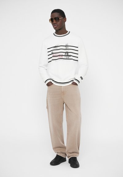 Λευκό φούτερ με μαύρες οριζόντιες ρίγες και γραφικό τύπωμα· συνδυασμένο με μπεζ παντελόνι από κορντόρι και μαύρα αθλητικά παπούτσια. Φοράει μαύρα γυαλιά ηλίου.