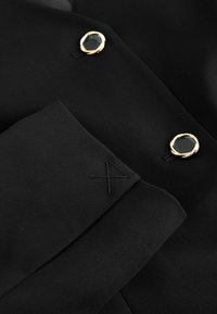 Sort stof blazer med to guld-accenterede knapper med mørke sten i midten. Inkluderer en foldet manchet med broderede detaljer.