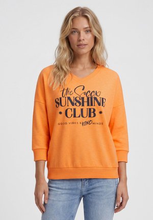 Blonde woman wearing an orange sweatshirt with black text reading "the soccx SUNSHINE CLUB Good vibes & kind minds" and blue jeans.