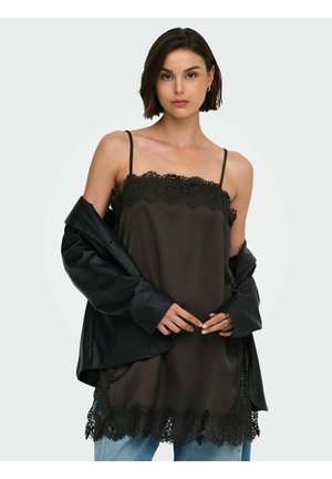 Femme portant un caraco noir orné de dentelle sur un jean avec une veste en cuir noire tombant des épaules, posant devant un fond clair.