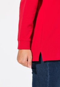 Maglietta rossa a maniche lunghe con vestibilità rilassata, caratterizzata da uno spacco laterale all'orlo e polsini a coste. Il materiale appare liscio e morbido.