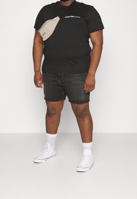 T-shirt nera a maniche corte con il logo bianco "Calvin Klein Jeans", abbinata a pantaloncini in denim scuro e sneakers alte bianche. Accentuata da una borsa a tracolla beige.
