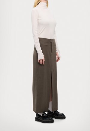 Kobieta ubrana w biały golf, brązową długą spódnicę z wysokim stanem i przednim rozcięciem, białe skarpetki oraz czarne masywne buty na jednolitym tle.