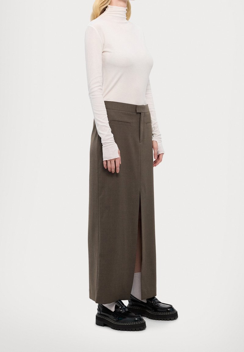 Femme portant un pull à col roulé blanc, une longue jupe marron taille haute avec une fente à l'avant, des chaussettes blanches et des chaussures épaisses noires sur un fond uni.