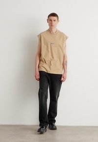 Beige bezrukávový bavlněný top, kulatý výstřih, logo na hrudi. Černé džíny z denimu s rovný fit. Černé boty, minimalistické pozadí.