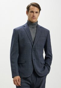 Giacca a quadri navy realizzata in tessuto testurizzato, dotata di due bottoni, rever a punta e tasche. Indossata sopra un maglione grigio a collo alto.