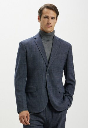 Giacca a quadri navy realizzata in tessuto testurizzato, dotata di due bottoni, rever a punta e tasche. Indossata sopra un maglione grigio a collo alto.