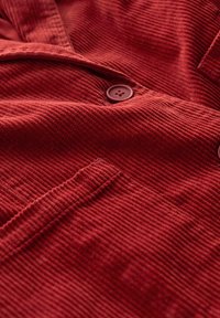 Tessuto di corduroy rosso con superficie testurizzata, due bottoni e taschino sul petto, che enfatizza il suo motivo a coste e il colore ricco.