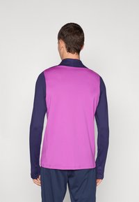 Camisola atlética de mangas longas com um corpo roxo e mangas azul-escuro, feita de um tecido liso e elástico com um design simples.