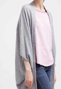 Ljusgrå, översized cardigan med öppen front och vida ärmar, bärs över en mjuk rosa T-shirt, kombinerad med blå denimjeans.