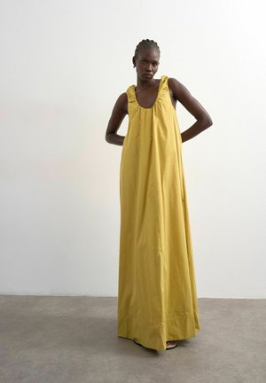 Femme aux cheveux tressés, debout les mains derrière le dos, portant une longue robe sans manches jaune moutarde devant un mur uni.
