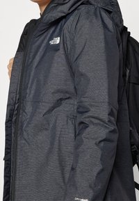 Vattentålig svart jacka med texturerad finish, dragkedja, justerbar huva och logotyp på bröstet. Har en lätt design.