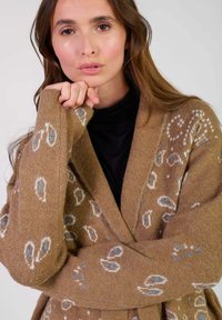 Cardigan en maille marron avec un motif paisley en blanc et bleu, col châle et manches larges, fabriqué en tissu texturé doux.