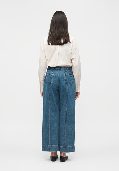 Γυναίκα με μακριά σκούρα μαλλιά, που φοράει ανοιχτό μπεζ μπλούζα με μακριά μανίκια, φαρδιά μπλε τζιν και μαύρα παπούτσια, γυρισμένη με την πλάτη προς το φακό σε μονόχρωμο φόντο.