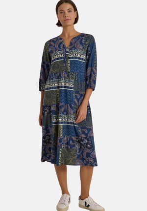 Femme debout portant une robe midi patchwork bleue et verte avec manches trois-quarts et baskets blanches.