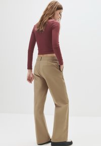 Braune weite Hosen mit seitlichen Taschen, kombiniert mit einem figurbetonten weinrotfarbenen Langarmoberteil. Die glatte Textur und das maßgeschneiderte Design sind sichtbar.