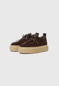 Baskets en daim marron avec une semelle épaisse beige, bout rond et lacets. Présente un col rembourré et une semelle texturée pour une meilleure adhérence.