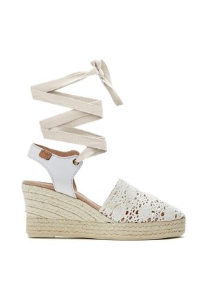 Alpargata blanca con cuña, parte delantera de ganchillo, lazos para atar en el tobillo y tacón con textura de cuerda sobre un fondo blanco.