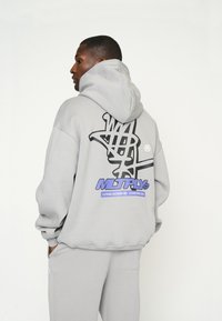 Grauer Kapuzenpullover mit großem schwarzem Grafikdesign auf der Rückseite und lila Logo-Akzenten. Geschmeidige Textur und lockere Passform.