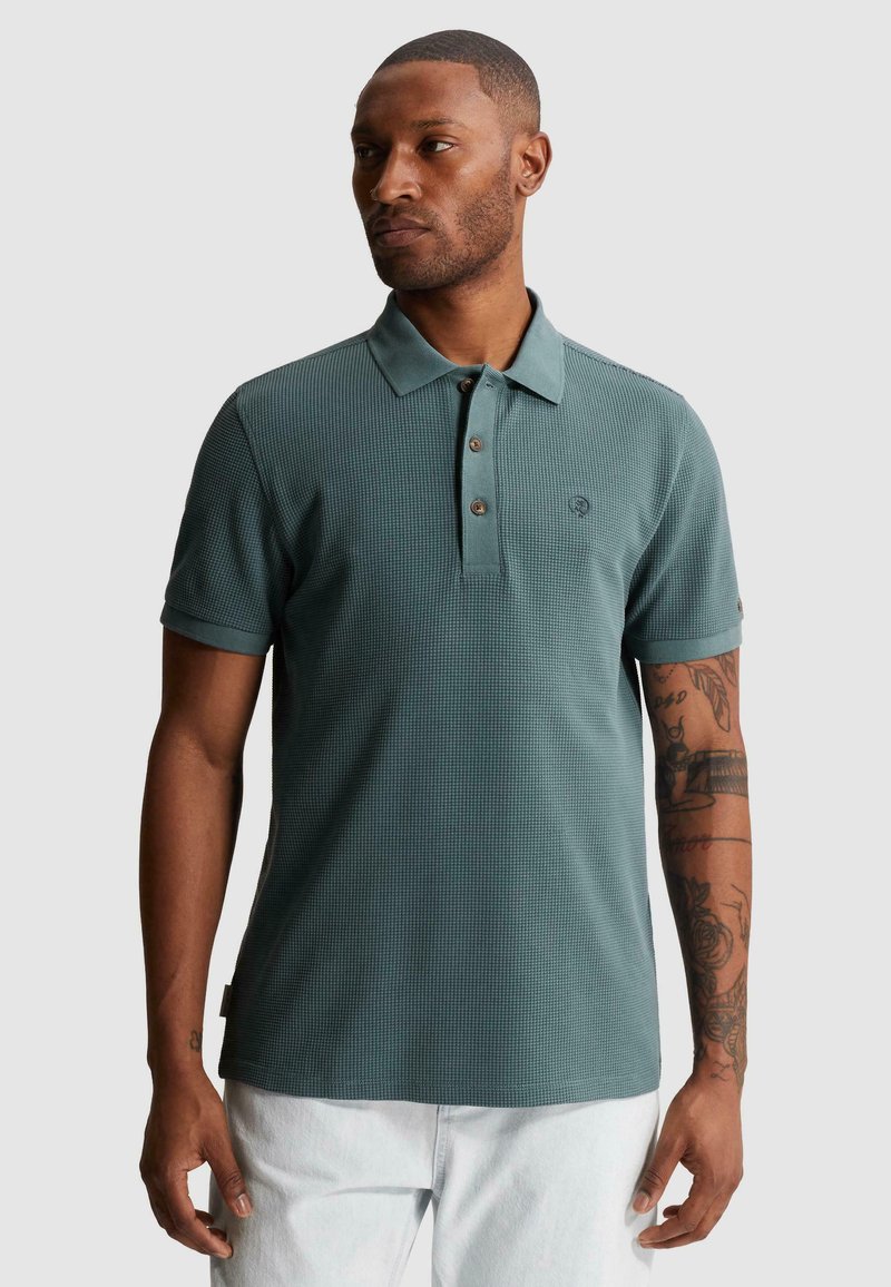 Homme portant un polo texturé de couleur sarcelle avec des boutons et un pantalon blanc, debout devant un fond clair uni.