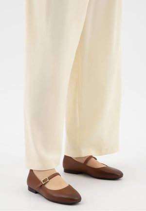 Chaussures plates Mary Jane en cuir marron avec brides à boucle portées avec un pantalon à jambes larges crème sur fond blanc.