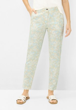 Lyseblå og beige blomstret print slim-fit bukser båret med hvide slidesandaler, model stående med den ene hånd i lommen.