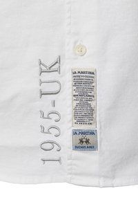 Bílá košile s vyšitým nápisem "1955-UK" v šedé barvě. Obsahuje dvě etikety: jednu modrou s textem, druhou s logem. Hladká textura látky.