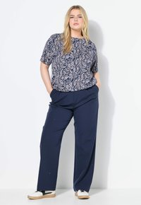Donna bionda in piedi con le mani nelle tasche dei pantaloni blu, indossa un top a maniche corte blu e bianco con un motivo e sneaker bianche con dettagli neri.